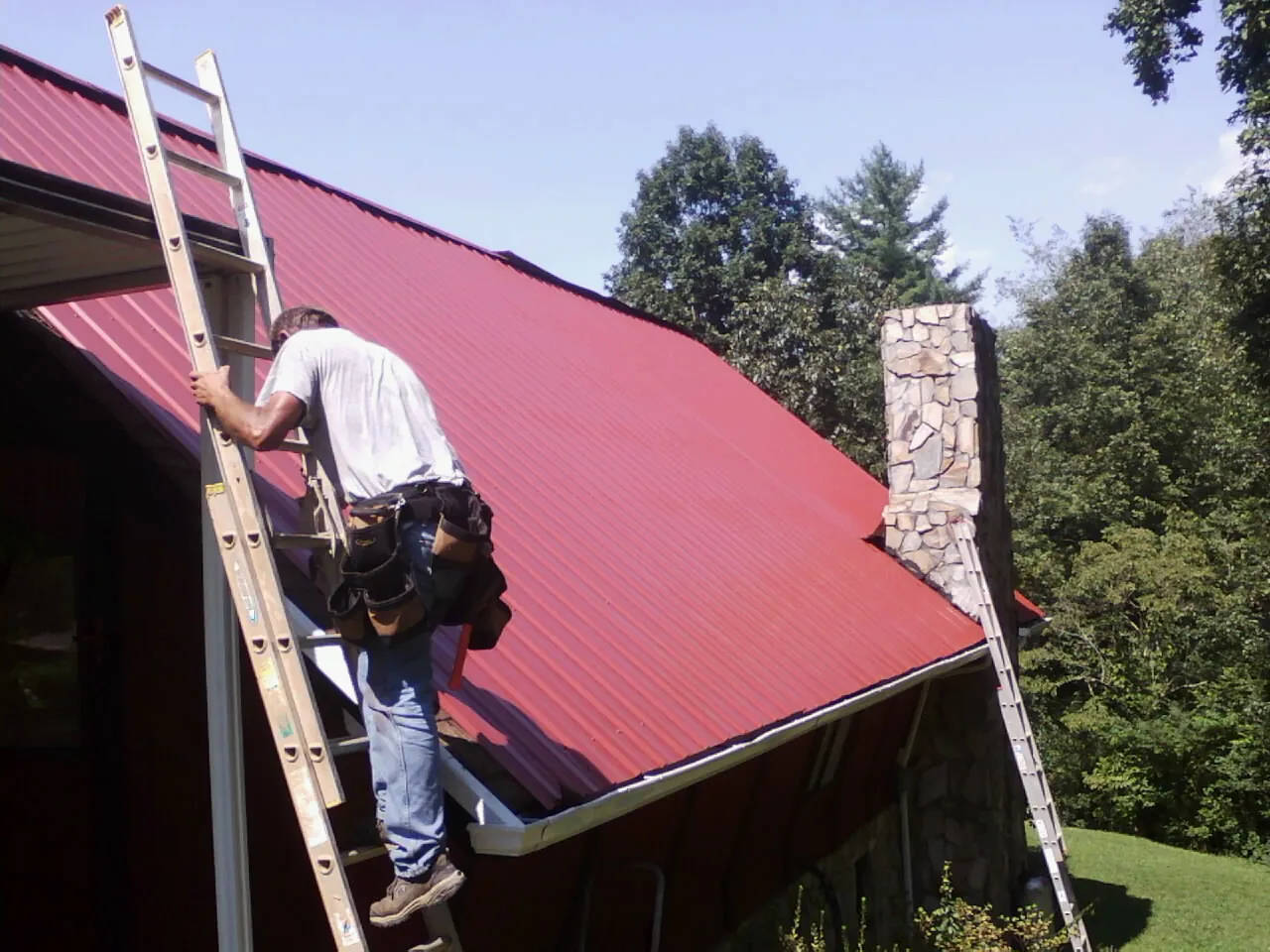 Roofer on a ladder beside a red metal roof and stone chimney — Soffit & Fascia Repair by Roofing Brothers in Owego